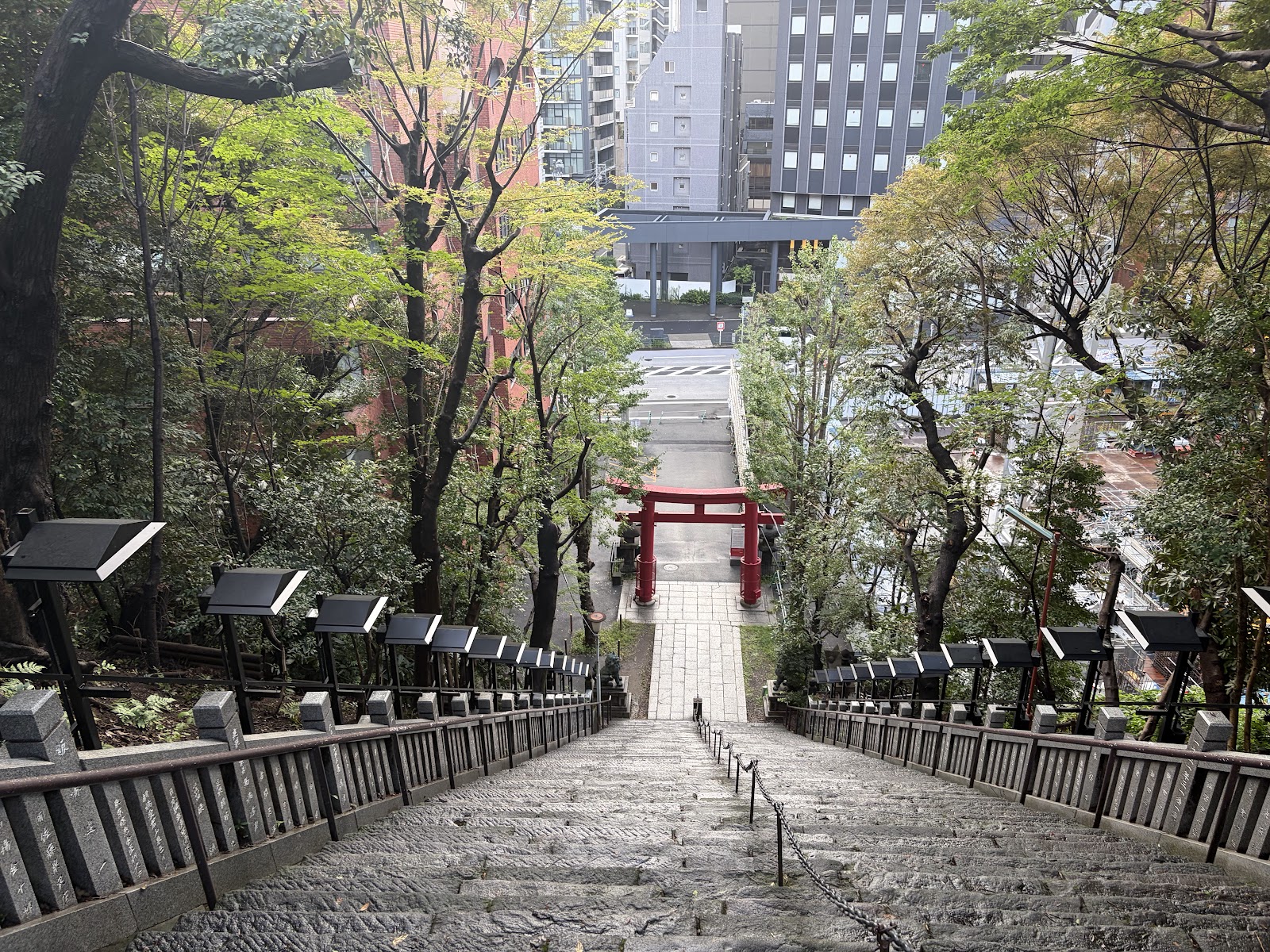 上から見下ろした石段。下に朱の鳥居、その先に道路と高層ビル、両側に木々と灯籠。