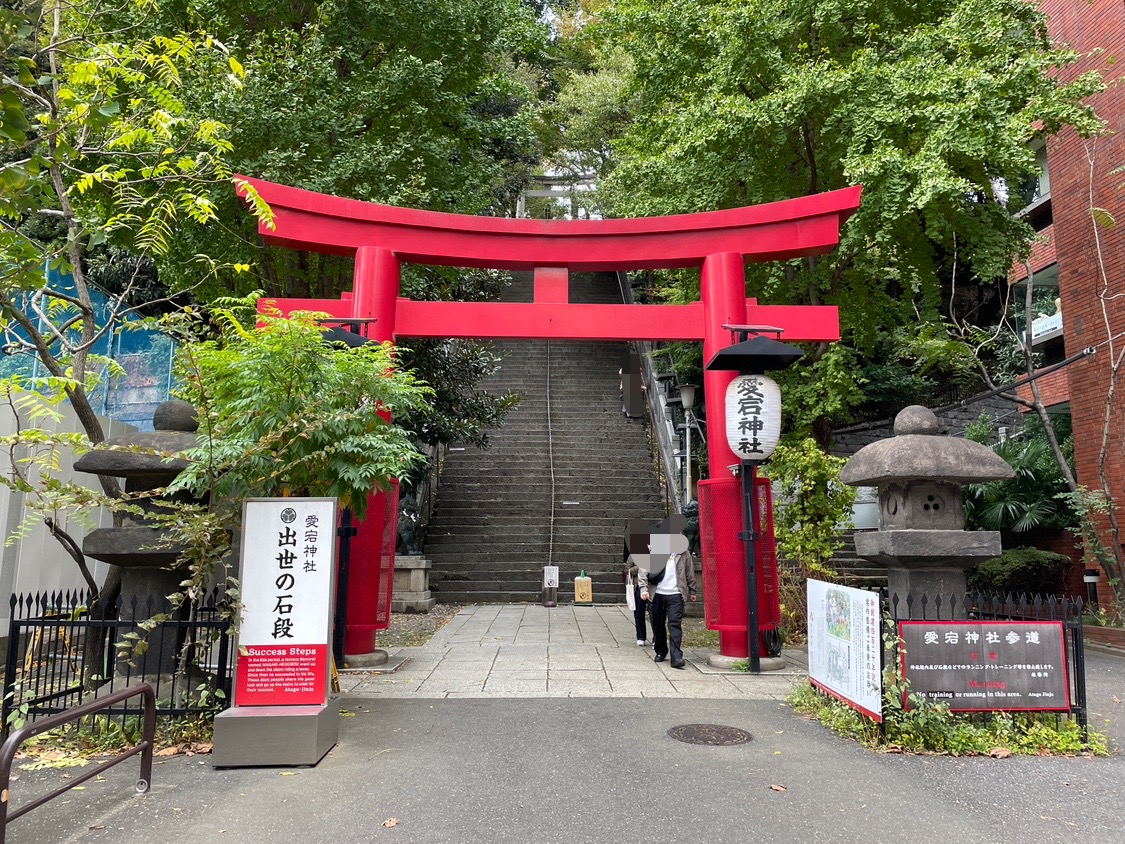 愛宕神社の朱の鳥居と、その奥に続く石段。左に出世の石段の立て札、右に提灯と石灯籠。
